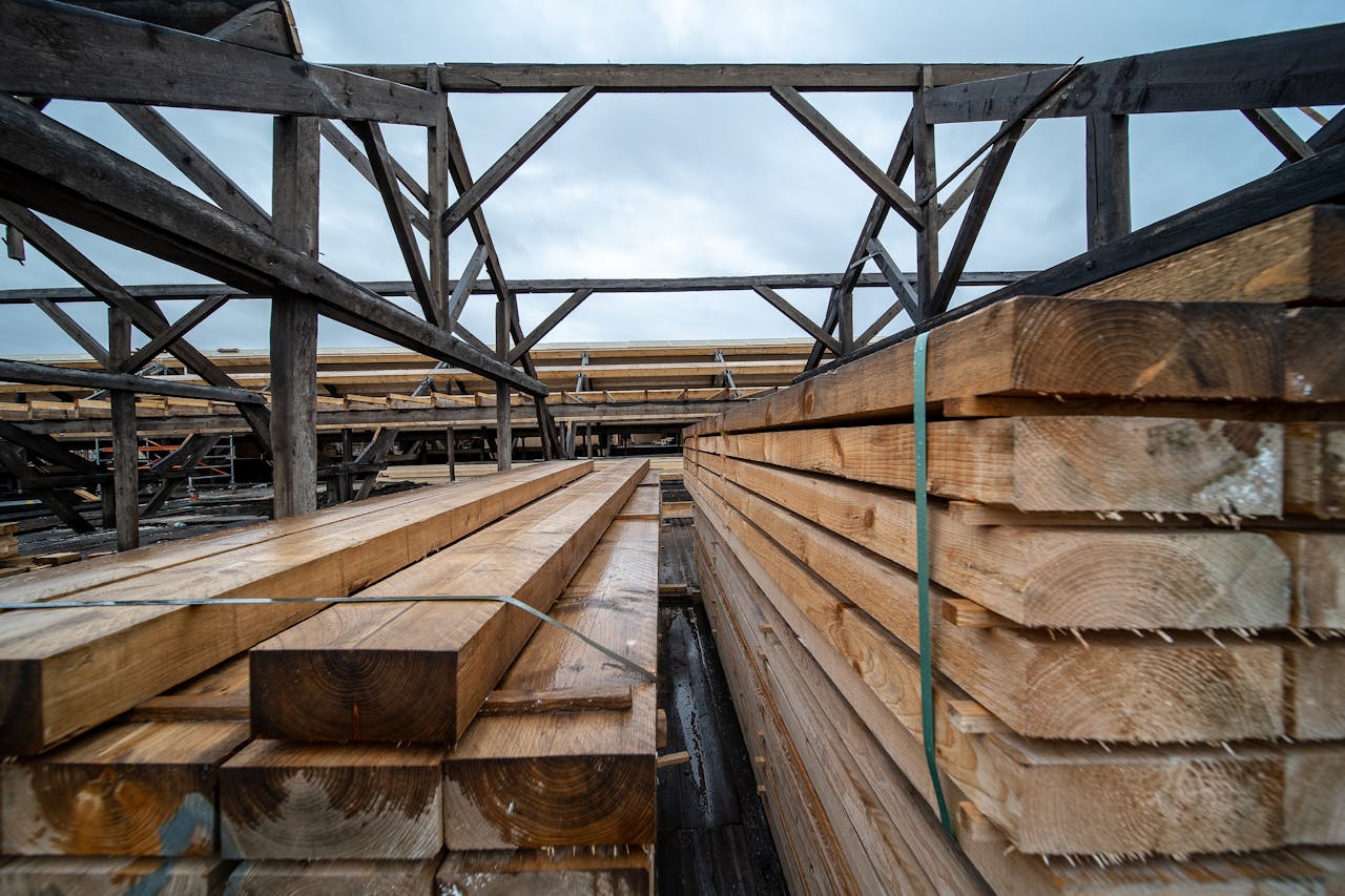 Stacked timber planks in an outdoor industrial storage area under a cloudy sky.